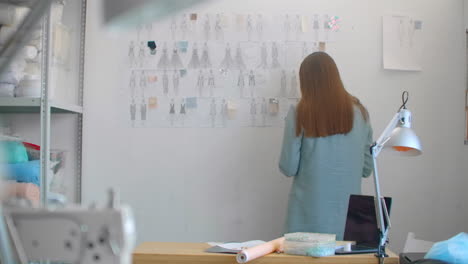 Young-fashion-designer-standing-with-back-to-camera-looking-at-drawings-sketches-hanging-on-wall.-Woman-is-thinking-about-new-trendy-clothing-collection.